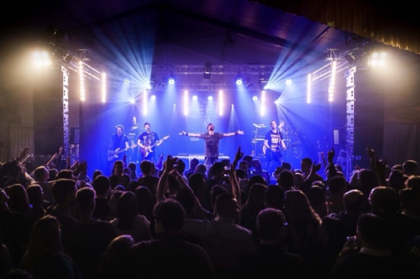 Band auf einer Bühne im blauen Scheinwerferlicht vor dichtem Publikum mit erhobenen Armen