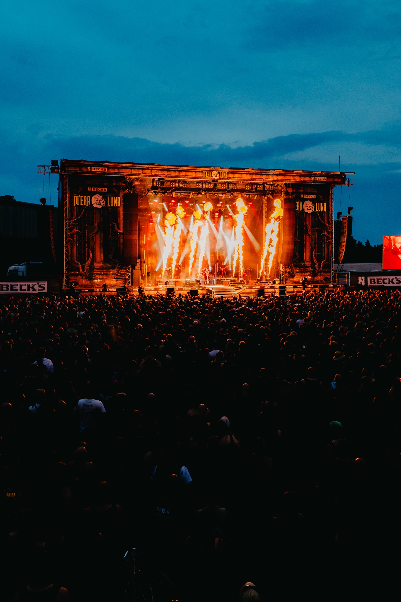 Festivalbühne mit Flammensäulen und Lichtstrahlen vor großem Publikum bei Dämmerung