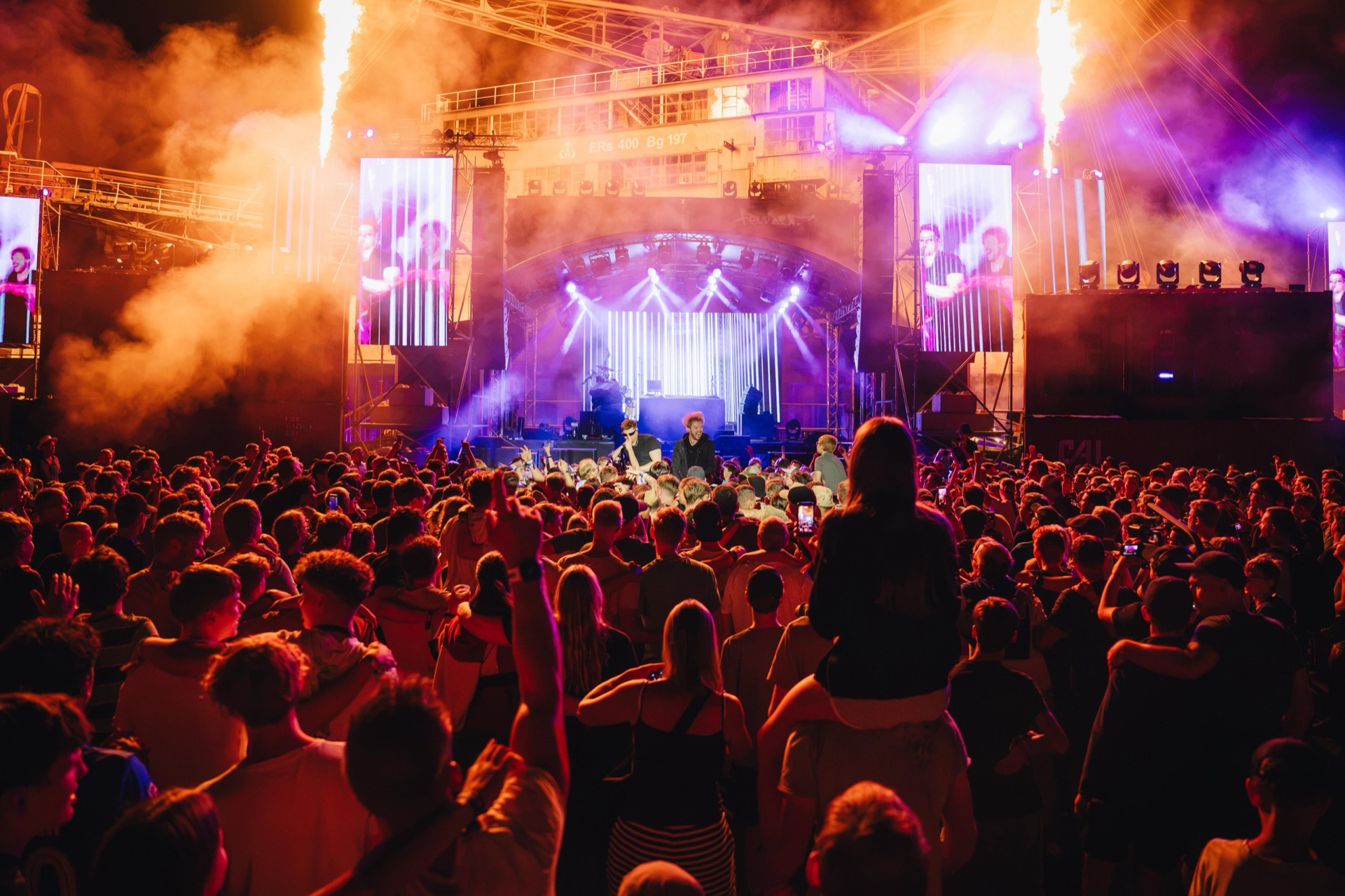 Festivalbühne mit seitlichen Flammenbalken und Bildflächen, davor das Publikum