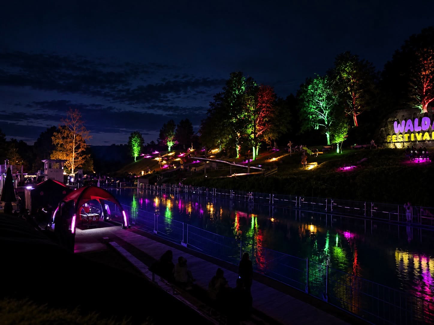 Bunt angestrahlte Bäume spiegeln sich im Wasser, daneben der Schriftzug Waldfestival
